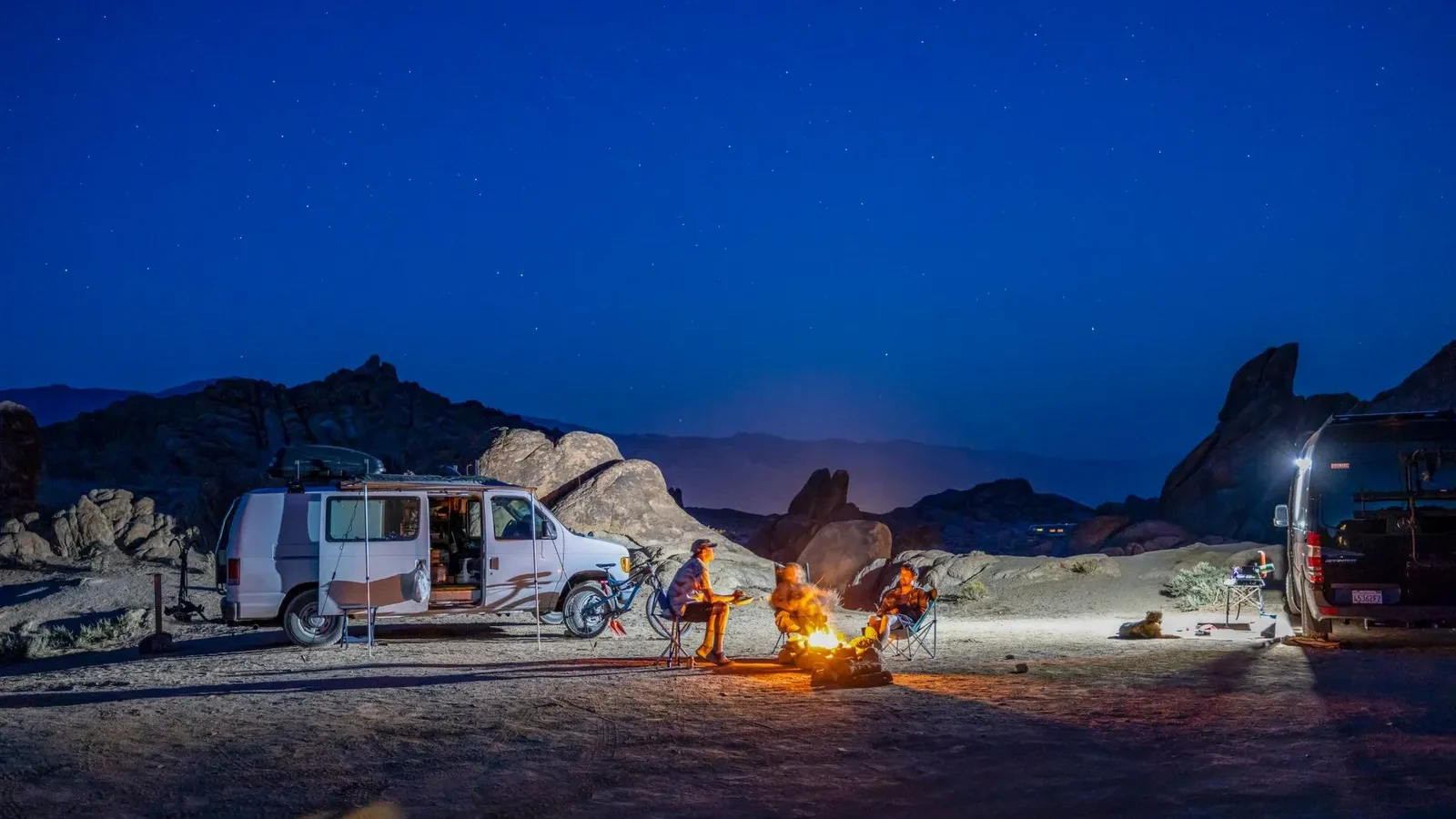 Camper van campsite at night with camp lights and a small fire under a starry sky