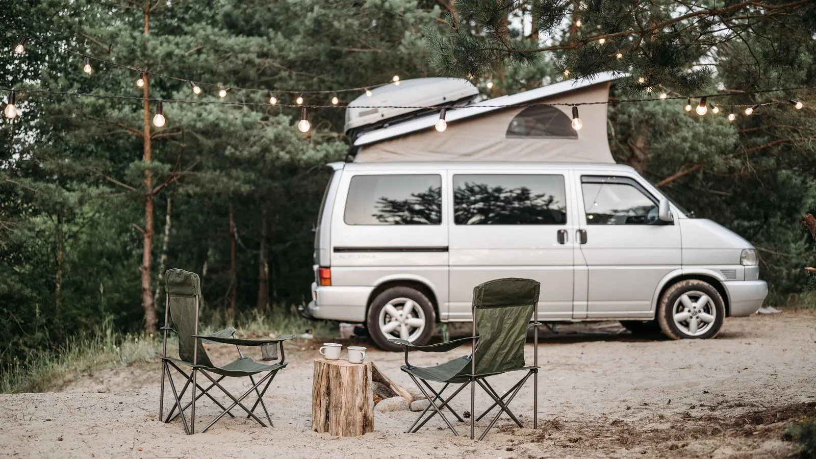 Camper van at a wooded campsite with chairs and string lights
