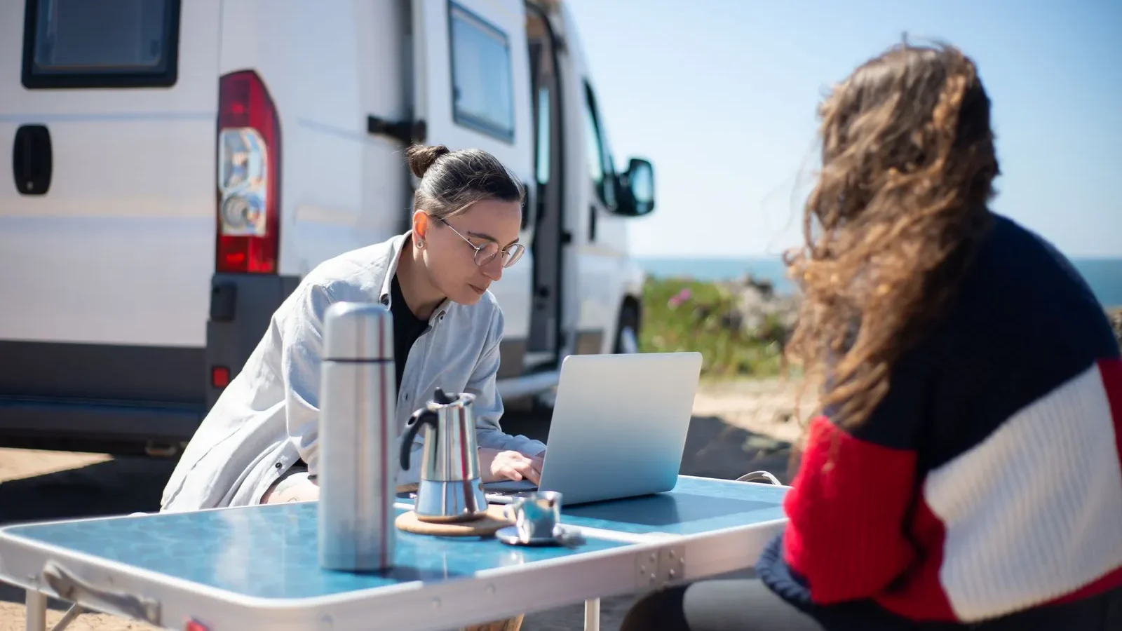 Two travelers working on a laptop at a folding table beside a camper van by the sea