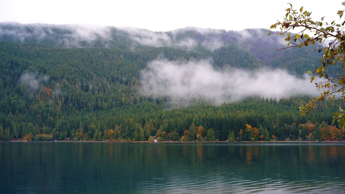 Calm lake with low clouds drifting over forested hills