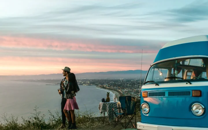 Couple beside a blue camper van at an ocean overlook at sunset