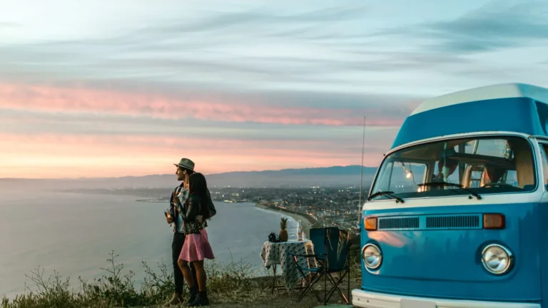 Couple beside a blue camper van at an ocean overlook at sunset