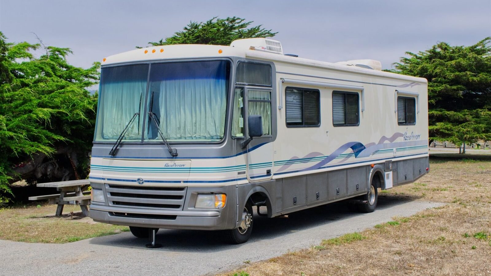 An older Class A motorhome parked on a gravel site among trees