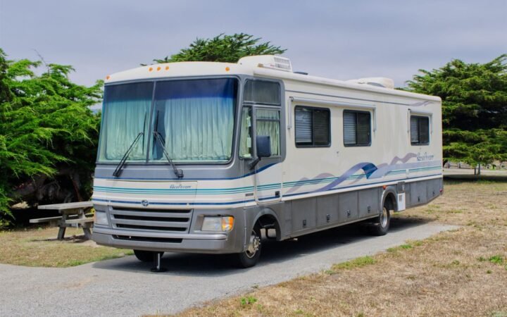 An older Class A motorhome parked on a gravel site among trees