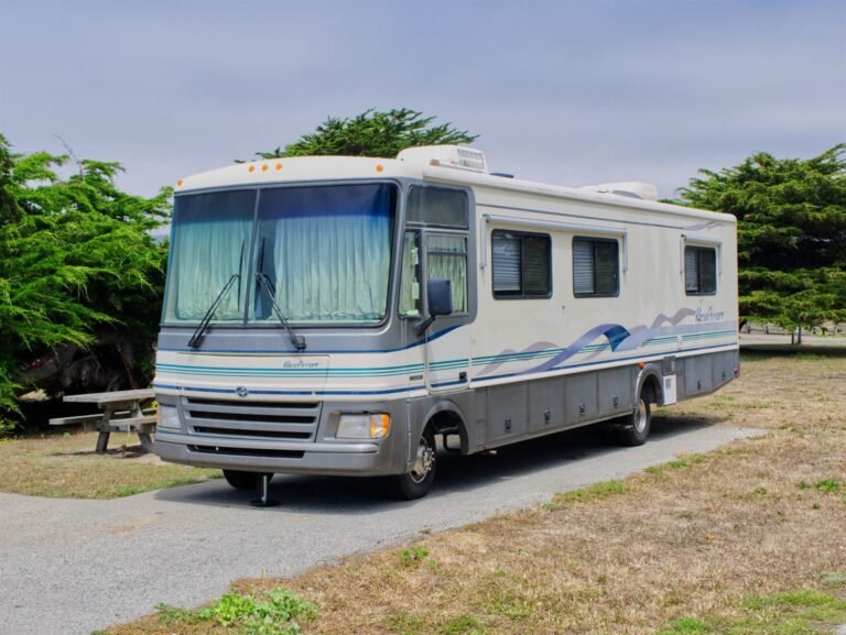 An older Class A motorhome parked on a gravel site among trees