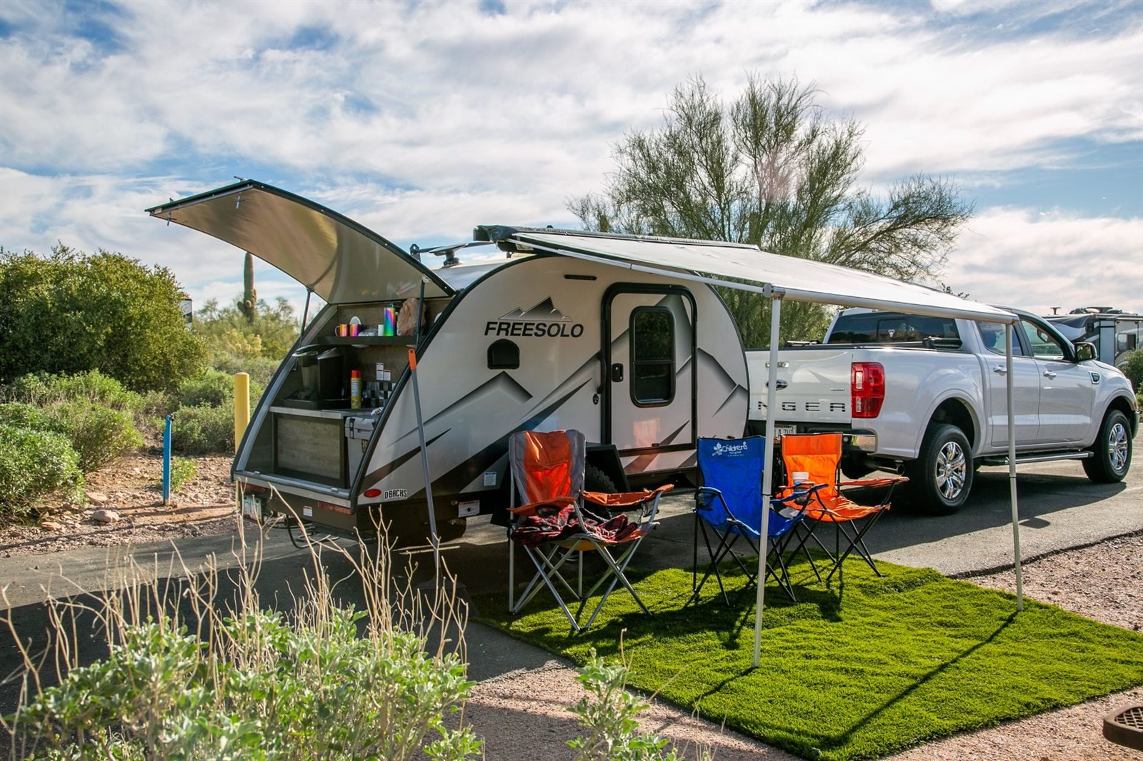A small modern travel trailer set up at a campsite behind a pickup truck, with an awning and camp chairs out